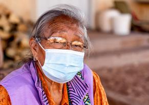 Older, female presenting person looks at camera wearing a mask and glasses