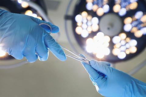 One blue-gloved hand is seen handing a pair of surgical scissors to another blue-gloved hand. A surgical light shines overhead.